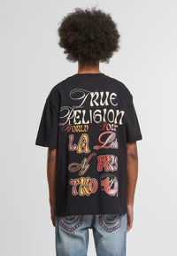 Camiseta negra de algodón con texto gráfico multicolor "True Religion World Tour" en la parte posterior. Manga corta, ajuste relajado.