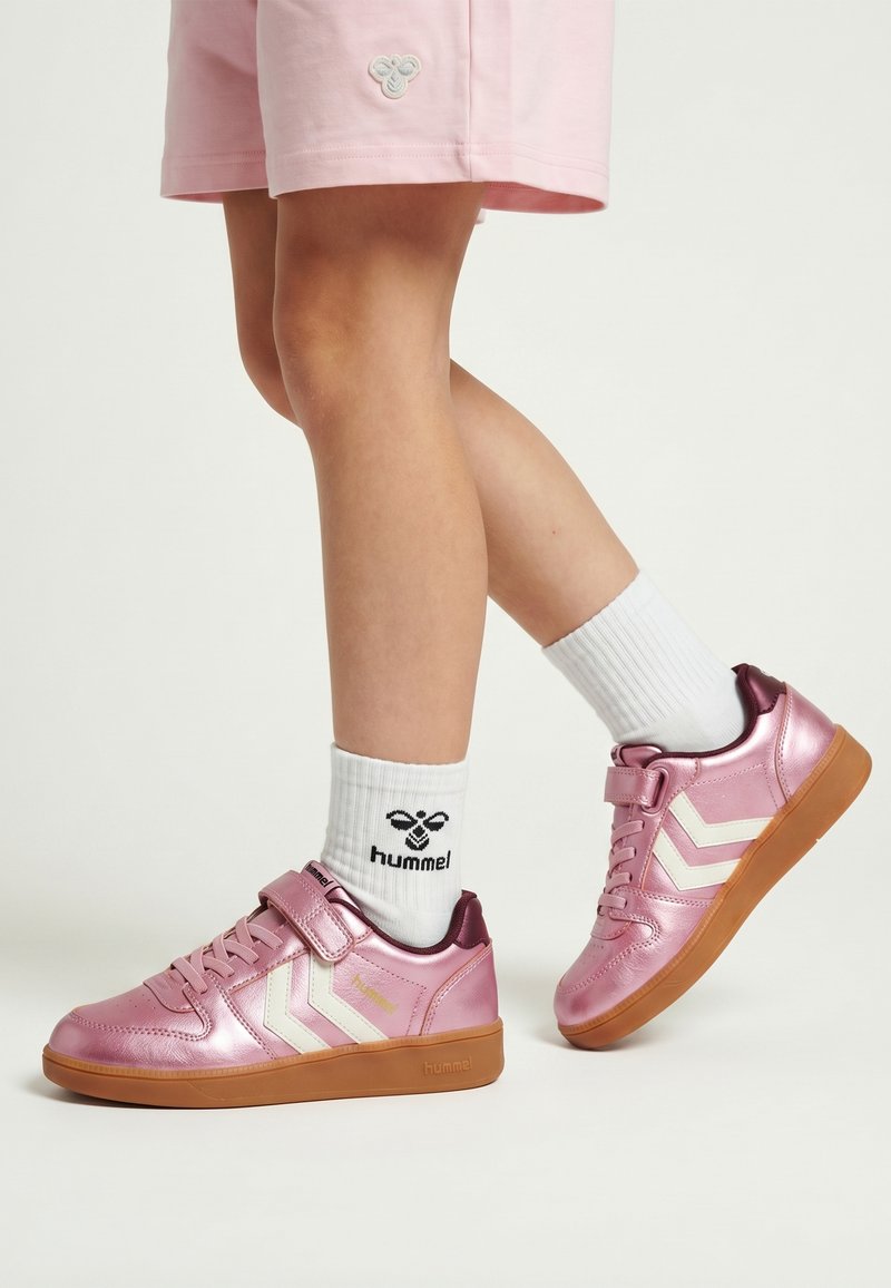 Barn iført pink metallic sneakers med hvide Chevron-striber, hvide Hummel-strømper og pink shorts mod en ensfarvet baggrund.