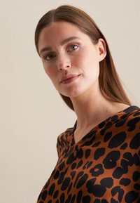 Blusa marrone in seta con stampa leopardata nera, design a V e texture liscia. Il tessuto ha una leggera lucentezza.