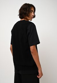 Pull en maille noir à manches courtes, avec une finition texturée et des bords côtelés. Le mannequin est montré de dos, portant un pantalon noir ample.