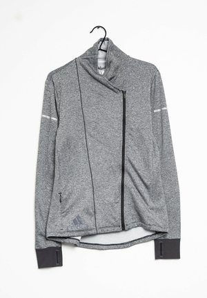 Veste Adidas grise à manches longues avec fermeture éclair, col montant, fermeture éclair noire asymétrique, trous pour les pouces aux poignets et logo près de la poche gauche, accrochée à un cintre noir.