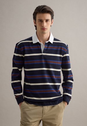 Jonge man draagt een rugbyshirt met lange mouwen in marineblauw, wit en bruin gestreept, met een witte kraag en beige broek, staand met zijn handen in zijn zakken.