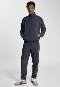 Marinblå träningsoverall i lättviktsmaterial med vita pipingsdetaljer, bestående av en jacka med dragkedja och byxor med elastisk midja. Sneakers med grå och blå accenter.