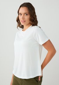 Vrouw met golvend bruin haar, gekleed in een eenvoudige witte T-shirt met korte mouwen en olijfgroene broek, staande met één hand in de zak.