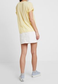 Žlutý top s krátkým rukávem a volány na manžetách, doplněný bílou sukní. Modelka má na sobě světle modré platformové tenisky a šedé ponožky.