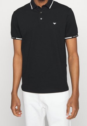 Mann trägt schwarzes kurzärmeliges Poloshirt mit weißem Besatz und Logo auf Brust und Ärmeln, kombiniert mit weißen Hosen, vor einfachem Hintergrund.