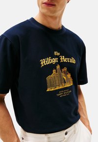 Camiseta azul marino con texto amarillo "The Hilfiger Herald" y un gráfico detallado de un horizonte urbano, combinada con pantalones blancos.