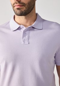 Polo de culoare mov deschis, dintr-un material texturat, cu un guler clasic și două butoane la gât. Mâneci scurte.