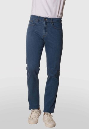 Persona que lleva vaqueros azules de pierna recta y zapatillas blancas, de pie frente a un fondo claro y liso.