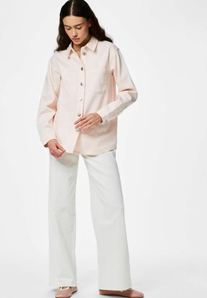 Jeune femme boutonnant une chemise rose pâle, portant un pantalon blanc à jambes larges et des ballerines roses, debout devant un fond uni.