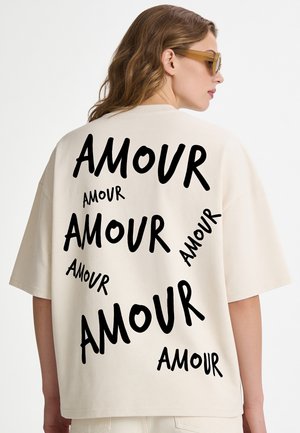 Femme portant un T-shirt beige oversize avec le mot "AMOUR" imprimé plusieurs fois en noir dans le dos, portant également des lunettes de soleil marron clair.