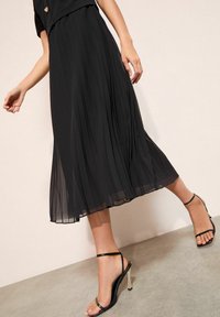 Fekete pliszírozott midi ruha, amely testhezálló felsőrésszel és áramló szoknyával rendelkezik, fekete pántos szandálokkal párosítva, melyek fémes sarokkal vannak ellátva.