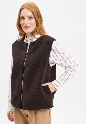 Bruine fleecedeken met een ritssluiting aan de voorkant, ronde halslijn en zijzakken, gelaagd over een witte lange mouwen shirt met rode strepen.
