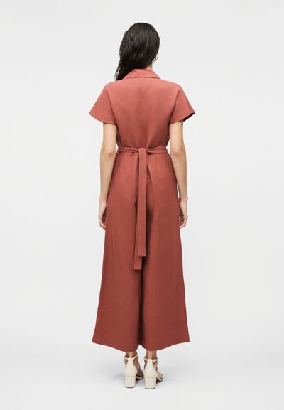 Γυναίκα με μακριά σκουρού χρώματος μαλλιά φοράει ολόσωμη φόρμα σκουρόχρωμου σκουριάς με κοντά μανίκια και ζώνη, και λευκά πέδιλα με τακούνι, στέκεται με την πλάτη στραμμένη σε απλό φόντο.