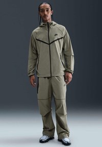 Olivgrön Nike-jacka och matchande byxor med svarta accenter. Jackan har huva och dragkedja; byxorna har lös passform och fickor.