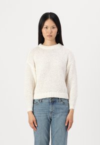 Cream-colored knitted sweater with a ribbed neckline and hem, featuring a loose fit and textured fabric. Paired with light blue jeans.