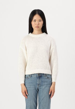 Pull en maille couleur crème avec un col et un ourlet côtelés, présentant une coupe ample et un tissu texturé. Associé à un jean bleu clair.