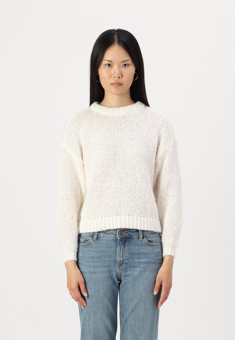 Cream-colored knitted sweater with a ribbed neckline and hem, featuring a loose fit and textured fabric. Paired with light blue jeans.