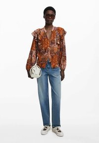 Oranje en paarse bloemenblouse met geruite mouwen, knopen aan de voorkant; gecombineerd met lichtblauwe spijkerbroek en witte sneakers. Houdt een witte tas vast.