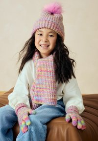 Cappello lavorato a maglia, sciarpa e guanti in tonalità di rosa, viola e blu; pompon soffice sul cappello; motivi testurizzati su tutti gli accessori. Camicia bianca e jeans.