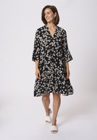 Robe noire à fleurs avec des manches courtes en forme de cloche, présentant un design à volants et de petites fleurs blanches. Portée avec des chaussures à talons ouverts blanches.