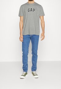 Camiseta de manga corta gris con el logo "GAP", jeans de denim azul y zapatillas verdes. Diseño simple y casual con un ajuste relajado.