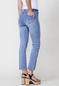 Svetlo modri jeans iz denima s prilegajočim krojem, petimi žepi in okrasnim šivanjem na zadnjih žepih. Kombinirani z črnimi sandali na peto.