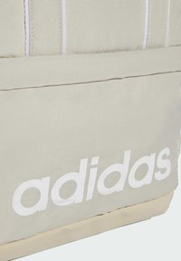 Bolso de lona beige con el logo blanco de "adidas", costuras en contraste y una forma estructurada con una superficie texturizada.