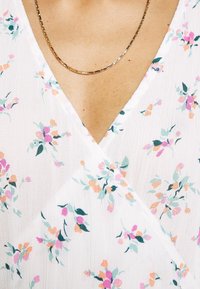 Gros plan d'une personne portant un délicat collier chaîne en or et une robe blanche à motifs floraux avec de petites fleurs roses, orange et vertes.