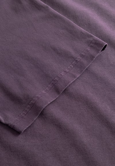 Lilla bomullsstoff med en glatt tekstur, med en halslinning og ermekant. Sømmene er synlige langs kantene.