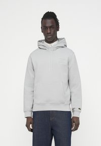 Sweat à capuche gris clair en matière douce, doté d'une poche avant, d'une capuche avec cordon de serrage et d'un petit écusson logo. Associé à un jean bleu foncé.