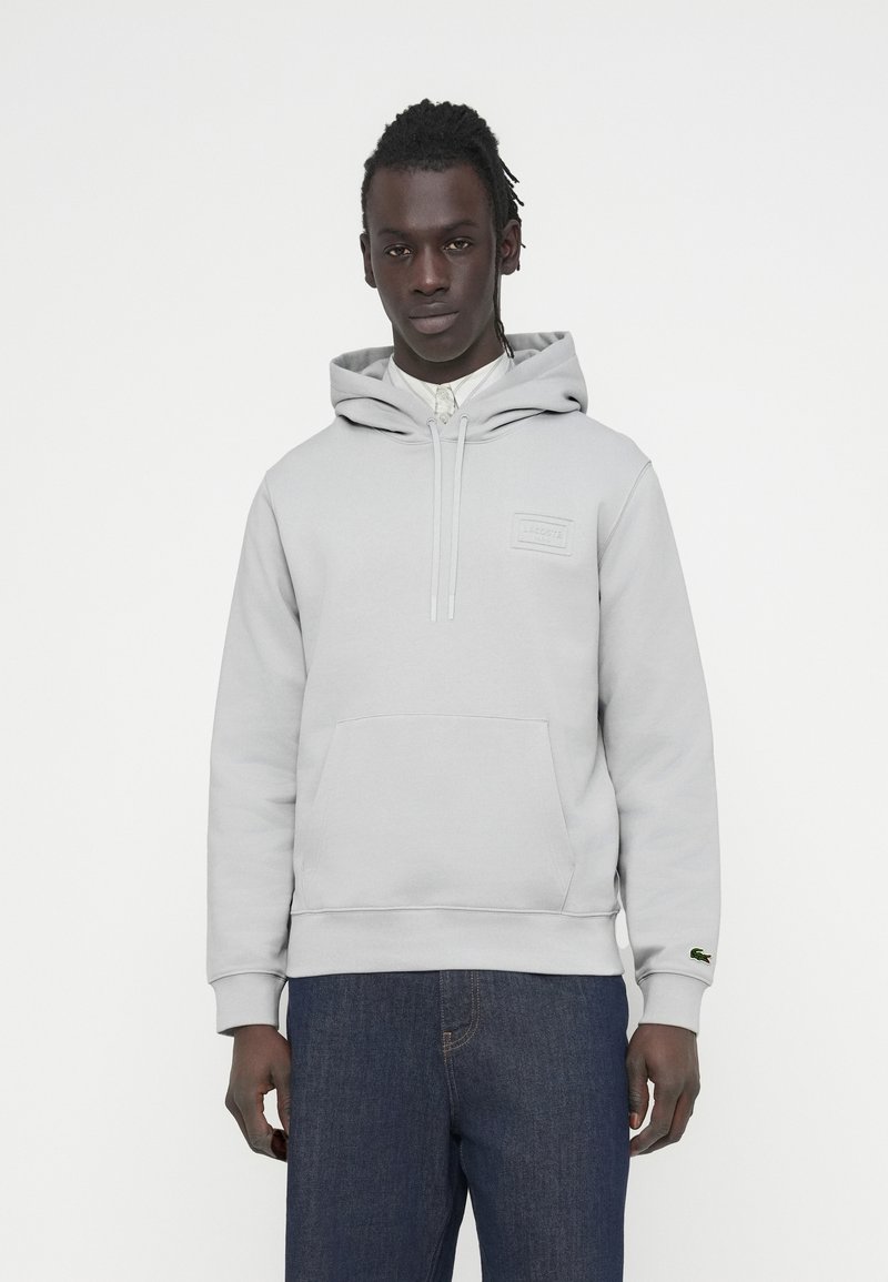 Sweat à capuche gris clair en matière douce, doté d'une poche avant, d'une capuche avec cordon de serrage et d'un petit écusson logo. Associé à un jean bleu foncé.