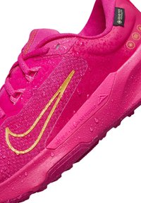 Zapatilla impermeable rosa con parte superior texturizada, logo en acento dorado y apariencia húmeda por gotas de agua en la superficie.