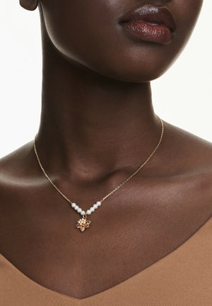 Nahaufnahme einer Frau, die eine goldene Halskette mit weißen Perlen und einem floralen Anhänger mit rosa und gelben Edelsteinen trägt.