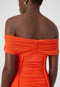 Πορτοκαλί φόρεμα off-the-shoulder από ραμμένο ύφασμα, με υφή και εφαρμοστό σιλhouette. Διακριτικά ασημένια σκουλαρίκια ορατά.