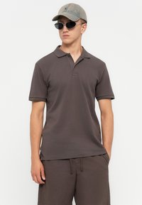 Ung man iklädd mörkgrå pikétröja och matchande shorts, med en grå keps och svarta solglasögon, som står mot en vit bakgrund.