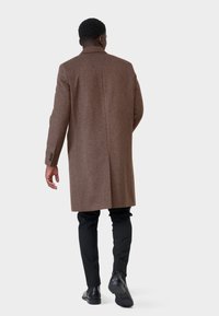 Bruine wollen overjas met een op maat gemaakte snit, enkele split en notch revers. Gecombineerd met zwarte broek en schoenen, gezien vanuit de achterkant.