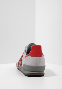 Suedefärgade grå sneakers med röda accenter, som har tre sidoremsor, ett vadderat ankelskydd och en texturerad gummisula.