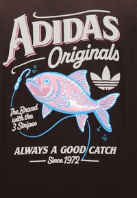 Λογότυπο Adidas Originals και τριφύλλι δίπλα σε ένα ροζ και μπλε ψάρι στο αγκίστρι, με το κείμενο "Πάντα μια καλή ψαριά από το 1972."