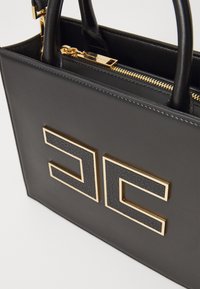 Sac à main en cuir noir avec matériel doré et un accent logo texturé. Dispose de doubles poignées et d'une fermeture éclair sur le dessus.