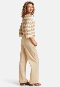 Beige gebreide top met een chevronpatroon en brede mouwen, gecombineerd met lichtbeige broeken. Model toont een zijaanzicht en draagt sandalen.