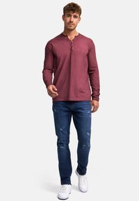 Rotes Langarm-Henley-Shirt mit knöpfbarem Ausschnitt, kombiniert mit gefleckten blauen Jeans und weißen Sneakers. Lässiger Schnitt und Design.