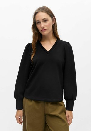 Frau mit langen braunen Haaren, die ein schwarzes langärmeliges V-Neck-Oberteil und olivgrüne Hosen trägt, steht vor einem schlichten weißen Hintergrund.
