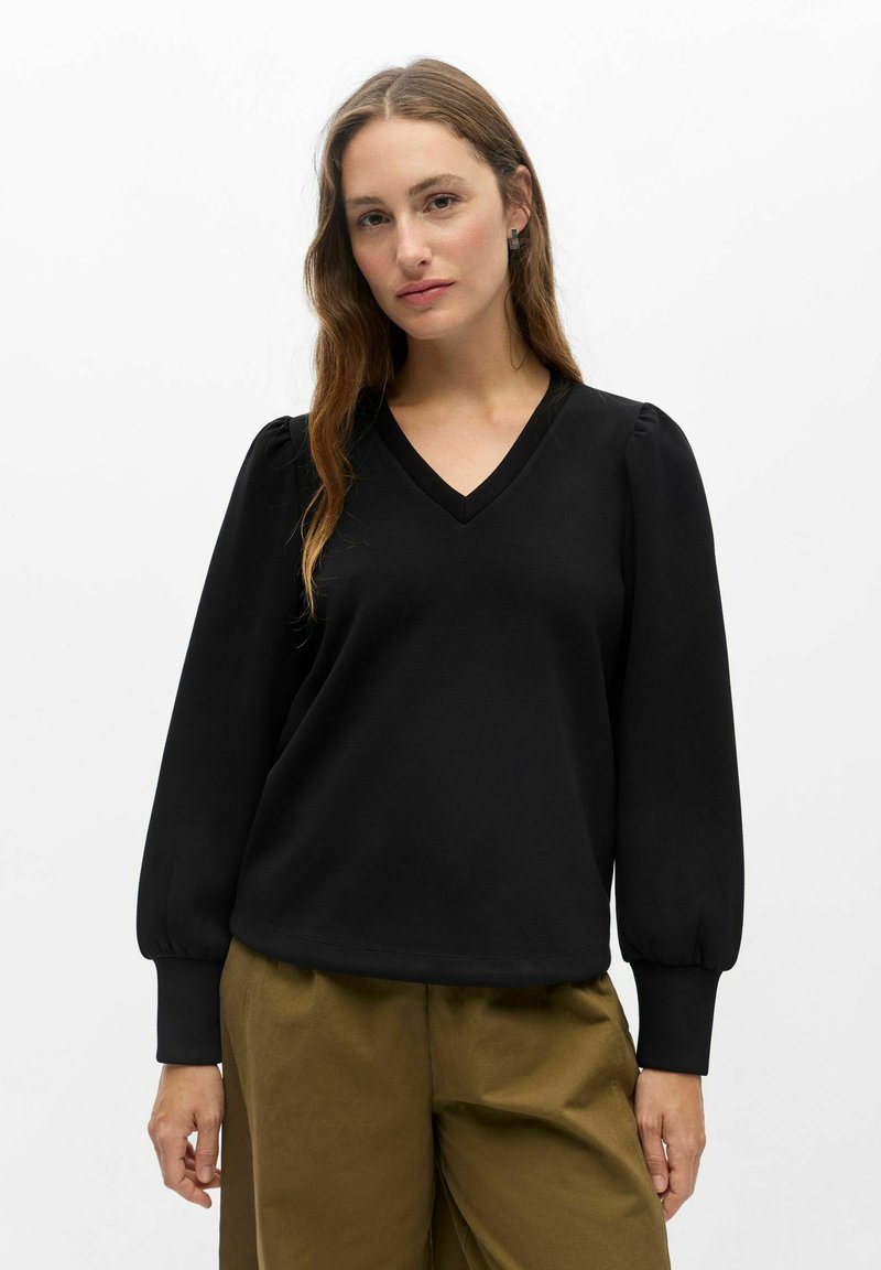 Femme aux longs cheveux bruns portant un haut noir à manches longues avec un col en V et un pantalon vert olive, debout devant un fond blanc uni.