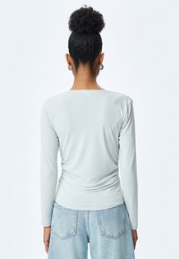Helles graues Langarmshirt mit einer gerafften Gestaltung an der Taille, featuring einem runden Ausschnitt und dekorativen Knöpfen an der Schulter.