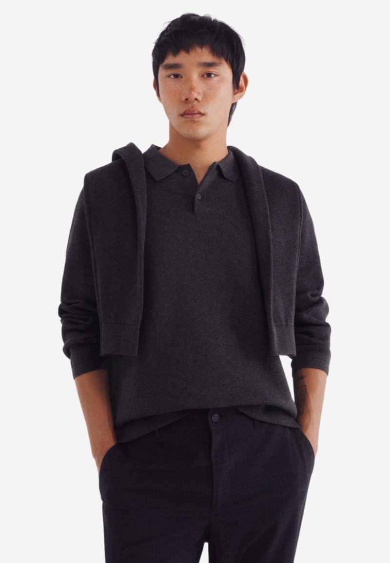 Polo a maniche lunghe grigio scuro con colletto e due bottoni, abbinato a un cardigan coordinato appoggiato sulle spalle.