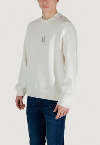 Weißes Crewneck-Sweatshirt aus weichem Stoff mit einem subtilen, goldenen Logo in der Mitte. Kombiniert mit dunkelblauen Jeans.