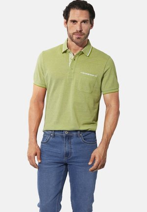 Mann trägt ein hellgrünes Kurzarm-Polohemd mit Brusttasche und blaue Jeans vor einem schlichten, hellen Hintergrund.