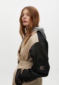 Beżowy i czarny płaszcz trench z paskiem w talii i długimi rękawami, z naszywką logo na lewym ramieniu oraz szarym kapturem pod spodem.