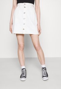 Gonna in denim bianco con bottoni frontali, vita aderente e orlo dritto. Abbinata a sneakers alte nere e calze bianche.